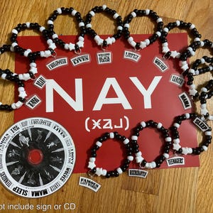May include: A collection of black and white beaded bracelets with charms featuring song titles and star-shaped beads. The bracelets are arranged around a red sign with the word "NAY" in white. A CD with a black and white design is also present.