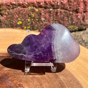 Puede incluir: Un cristal de amatista morado y blanco con forma de nube, sobre un soporte de acrílico transparente.