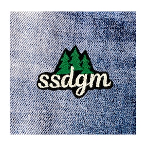 May include: A patch featuring three green pine trees atop a black mountain design, with the white text "ssdgym" below. The patch is sewn onto a blue denim fabric.