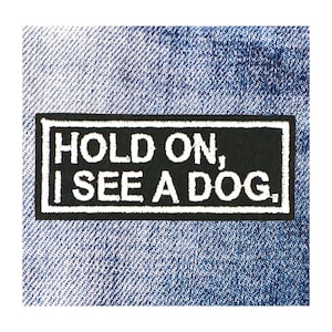 May include: A black rectangular patch with white text that reads "HOLD ON, I SEE A DOG." The patch is bordered in white and is set against a blue denim background.