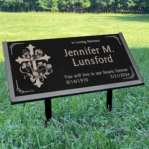 May include: Black memorial plaque with a decorative cross and the name "Jennifer M. Lunsford". Includes dates 8/14/1970 and 5/21/2024, and the inscription "You will live in our hearts forever". The plaque is set in a grassy area.