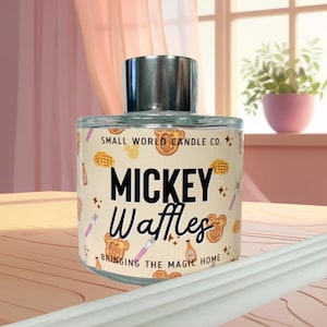 May include: A clear glass candle jar with a silver lid, labelled "Mickey Waffles." The label features waffle and Mickey Mouse head illustrations, along with the text "Bringing the Magic Home."