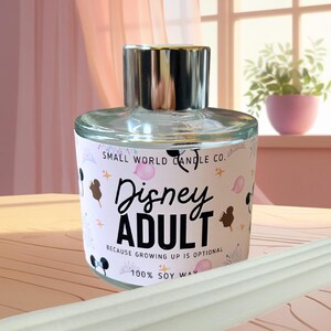 May include: A glass candle jar with a silver lid. The label says "SMALL WORLD CANDLE CO." and "Disney ADULT BECAUSE GROWING UP IS OPTIONAL 100% SOY WAX." The label has pink and black accents.