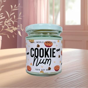 May include: A glass jar candle with a pale blue lid. The label reads "COOKIE Num" with cookie and star illustrations. The text "100% SOY WAX" is also on the label. The jar is on a wooden surface.