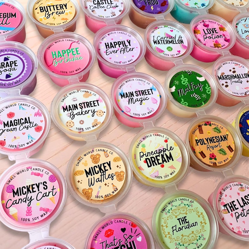 May include: A collection of colorful soy wax melts in small plastic containers. Each melt has a label with a unique name and design, such as "Mickey's Candy Cart" and "Happily Ever After." The melts are arranged on a light-colored surface.