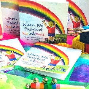 Puede incluir: Cuatro copias del libro infantil "When We Painted Rainbows" de Lisa Pearce. El libro presenta a un niño pintando un arcoíris con un pincel. Los libros están rodeados de juguetes y telas de colores.