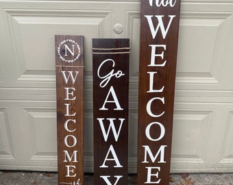 May include: Three wooden signs with white lettering. The first sign says "Welcome" with a small wreath and the letter "N" in the center. The second sign says "Go Away". The third sign says "Not Welcome".