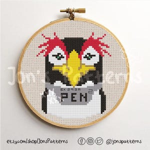 Pode incluir: Um aro de bordado de ponto cruz com um design de pinguim colorido. O pinguim está usando uma placa que diz "PEN" com um código de barras acima.