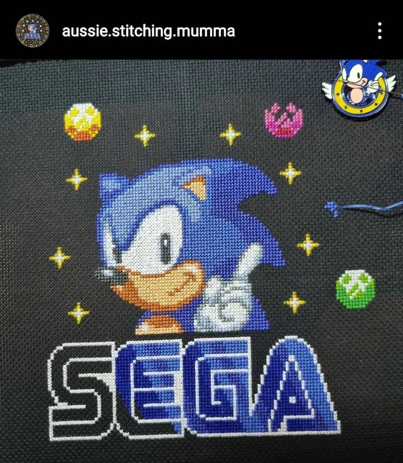 May include: Cross-stitch artwork featuring a blue Sonic the Hedgehog character with the word "SEGA" in blue and white letters on a black background. There are yellow and pink stars and rings around Sonic.