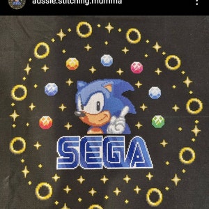 May include: A black fabric cross-stitch with a blue Sonic the Hedgehog character, the word "SEGA" in blue and white, and yellow rings and stars.