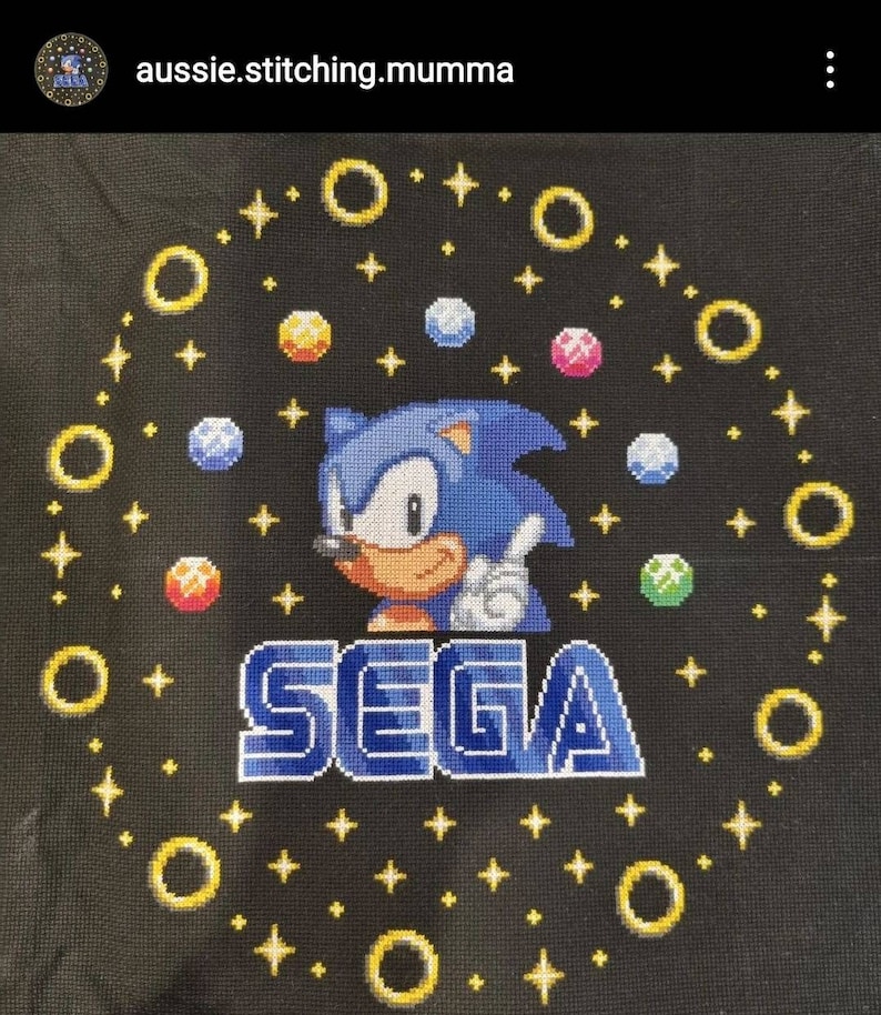 May include: A black fabric cross-stitch with a blue Sonic the Hedgehog character, the word "SEGA" in blue and white, and yellow rings and stars.
