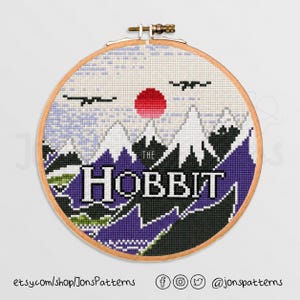 May include: A cross-stitch embroidery hoop featuring a landscape design with the text "THE HOBBIT." The design includes mountains, a red sun, and two birds. The color palette consists of white, purple, black, and green. The embroidery is set in a wooden hoop.