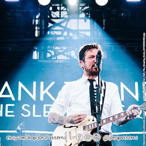 May include: A man with a beard and a black and white striped tie plays a white electric guitar on stage. The stage has a blue backdrop with the words "ANKON" and "HE SLE" in white text.