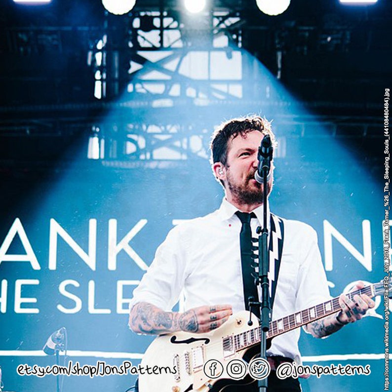 May include: A man with a beard and a black and white striped tie plays a white electric guitar on stage. The stage has a blue backdrop with the words "ANKON" and "HE SLE" in white text.