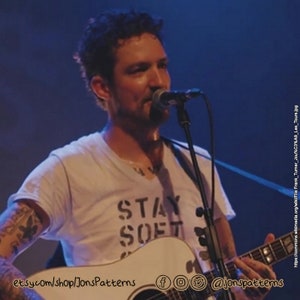 May include: A man with short brown hair and a beard is singing into a microphone while holding an acoustic guitar. He is wearing a white t-shirt with the words "Stay Soft" printed on it. The guitar is white and has a black pickguard. The man is performing on stage in front of a dark blue background.
