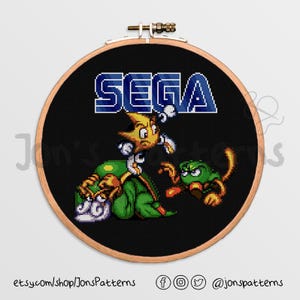 May include: A black and white cross-stitch pattern featuring the Sega logo and three pixelated characters. The characters are a yellow and blue star, a green and white creature, and a green and brown creature.