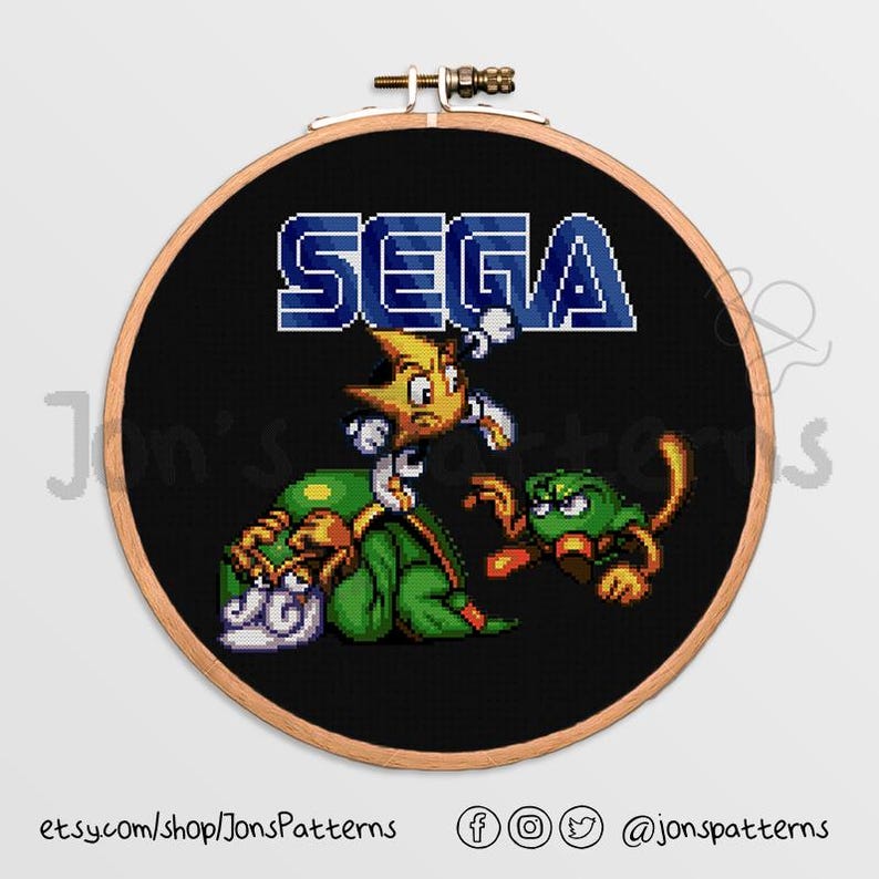 May include: A black and white cross-stitch pattern featuring the Sega logo and three pixelated characters. The characters are a yellow and blue star, a green and white creature, and a green and brown creature.