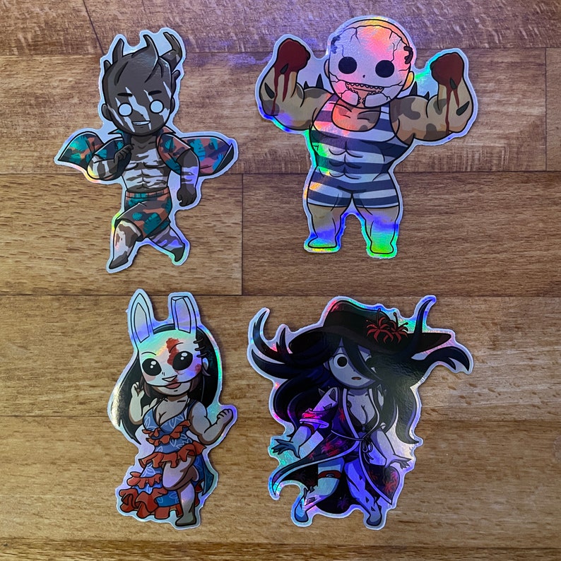 May include: Four iridescent holographic stickers featuring characters from a horror video game. The stickers are on a wood surface. The characters are a masked man with a striped shirt, a masked woman with a white shirt, a masked man with a green shirt, and a masked woman with a purple shirt.
