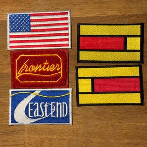 Può includere: Una collezione di toppe ricamate. Una toppa presenta la bandiera americana. Un'altra ha la parola "frontier" in giallo su sfondo rosso. Una terza toppa dice "East End" in bianco su sfondo blu. Due toppe hanno un design geometrico giallo e rosso.