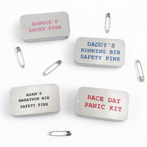 May include: Four small, rectangular silver tins with text in various colours and safety pins scattered around. The tins read: "Sophie's Lucky Pins," "Daddy's Running Bib Safety Pins," "Adam's Marathon Bib Safety Pins," and "Race Day Panic Kit."