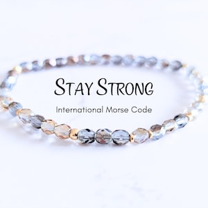 May include: A beaded bracelet with the words "Stay Strong" and "International Morse Code" in black text. The bracelet features clear and gold-colored beads. The beads are strung together to form a flexible band.