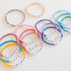 May include: A set of colorful beaded bracelets with positive affirmations, including "I am", "Fierce", "Loved", "Unique", "Enough", "Capable", "Strong", "Proud", "Grateful", "Kind", "Brave", "Calm", and "Worthy".