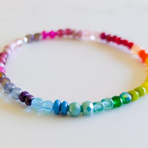 May include: A circular bracelet composed of small, multi-colored beads arranged in a gradient pattern. The beads feature various finishes, including translucent, opaque, and faceted textures, transitioning through shades of pink, red, orange, yellow, green, blue, and purple.