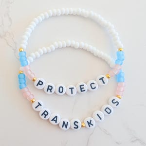 May include: Two beaded bracelets with white beads and gold accents. The top bracelet has blue and pink beads and spells out "PROTECT". The bottom bracelet has pink beads and spells out "TRANS KIDS".