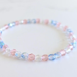 May include: A circular beaded bracelet featuring small, faceted, translucent beads in alternating shades of light pink, pale blue, and clear white. The beads are arranged in a repeating pattern, creating a delicate, reflective, and multi-colored appearance.