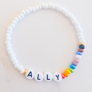 May include: White beaded bracelet with the name "ALLY" spelled out in black letters on white beads. The bracelet features a rainbow of colorful beads and gold accents.
