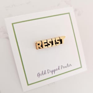 May include: Gold-dipped pewter pin with the word "RESIST" in bold, block letters. The pin is displayed on a white card with a green border, and the text "Gold Dipped Pewter" is printed below the pin.