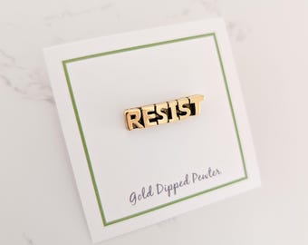 May include: Gold-dipped pewter pin with the word "RESIST" in bold, block letters. The pin is displayed on a white card with a green border, and the text "Gold Dipped Pewter" is printed below the pin.
