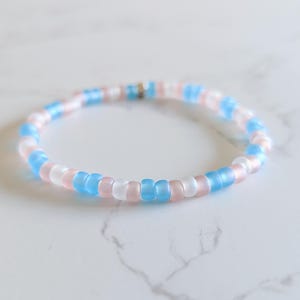 May include: A beaded bracelet featuring a pattern of light blue, pink, and white beads. The bracelet is made of small, round beads strung together. The bracelet is approximately 7 inches in circumference.