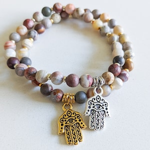May include: Double layered bracelet with brown, white, and gray agate beads and two silver hamsa hand charms. The hamsa charms have a gold eye in the center.
