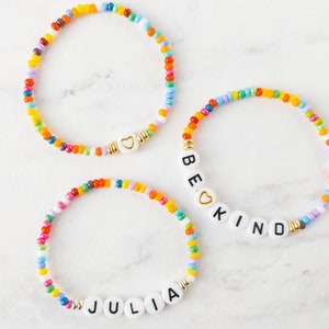 May include: Three colorful beaded bracelets with white letter beads spelling out "BE KIND" and "JULIA". Each bracelet has a small gold heart charm.
