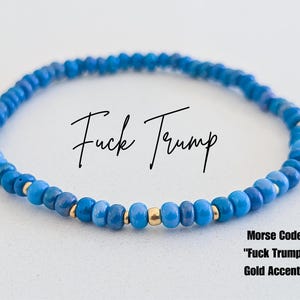 May include: A circular beaded bracelet featuring various shades of blue glass beads interspersed with small gold-colored metallic beads. The text "Fuck Trump" is written in the center, and the text "Morse Code "Fuck Trump" Gold Accents" is below.