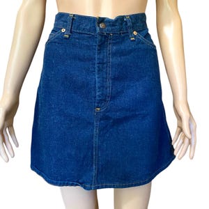 May include: A dark blue denim skirt with a classic design. The skirt features a button and zipper closure, front pockets, and yellow stitching. The skirt is a-line and has a vintage look.