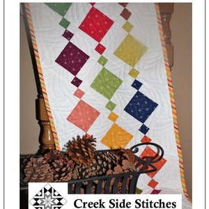 May include: A quilted table runner with a white background and colorful diamond patterns. The runner is 14.5 inches by 42.5 inches. The text "Beaded Charm" and "Creek Side Stitches" are visible. The runner is decorated with a variety of colors.