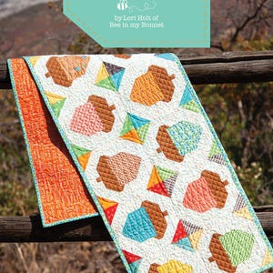 May include: A quilted table runner with an acorn and kite pattern. The runner features a white background with colorful acorns and kites. The runner is 15 inches by 65 inches. The text "ACORN LOVE" and "Table Runner Pattern" are visible.