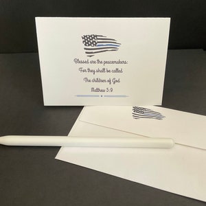 May include: A white card with a blue and black American flag with a thin blue line. The card has the text "Blessed are the peacemakers: For they shall be called The children of God Matthew 5:9".