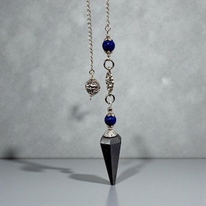 May include: A silver-toned pendulum with a faceted black crystal point. The pendulum features a chain with a decorative silver ball, blue beads, and ornate metalwork. The background is a neutral gray.