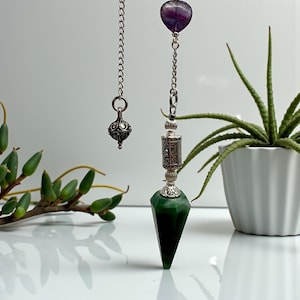 May include: A green gemstone pendulum with a silver chain and accents. The pendulum has a faceted, pointed shape. A purple heart-shaped stone is at the top of the chain. A silver decorative charm hangs from a separate chain.