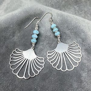 May include: A pair of silver earrings with a fan-shaped design. Each earring features a series of light blue beads suspended from a silver hook. The fan-shaped portion has a textured, matte finish.