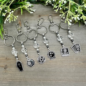 May include: Six silver keychains with clear crystal beads and black charms. The charms include a coffin, crescent moon, pentagram, crystal ball, book with the word "SPELLS" on the cover, and a deck of tarot cards.