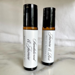 Puede incluir: Dos botellas de aceite de perfume roll-on con tapas negras y etiquetas blancas. Una está etiquetada como "Sandalwood & Jasmine", la otra "Jasmine & Rose". Las botellas son de vidrio color ámbar y las etiquetas tienen una escritura elegante.