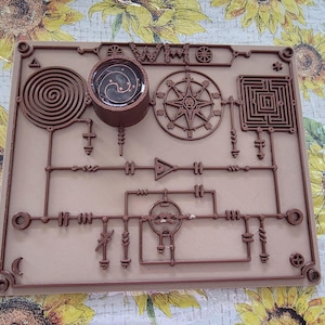 May include: A square, light brown board featuring a raised, dark brown circuit-like design. The pattern includes a spiral, a circular compass-like emblem, a square maze, and various geometric symbols connected by lines. A small, dark, circular container is attached to the top left.