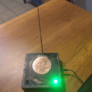 May include: A black wooden box with a copper-colored moon design on the top. A green LED light is on the front of the box. A silver antenna extends from the top of the box.