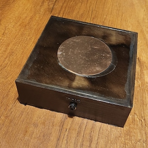 May include: A black wooden box with a copper-colored circular insert. The box has a small black plug on the front.