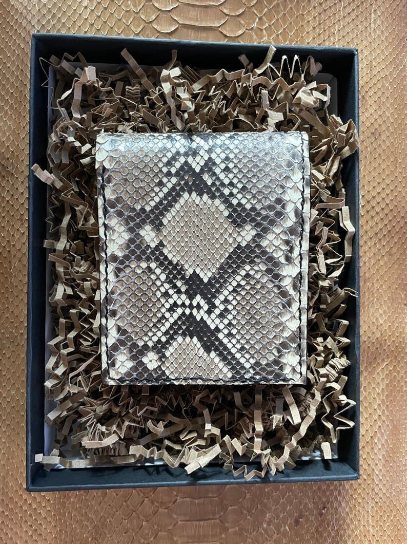 Pu&ograve; includere: Un portafoglio in pelle di serpente marrone e nero in una scatola regalo nera con carta triturata marrone.
