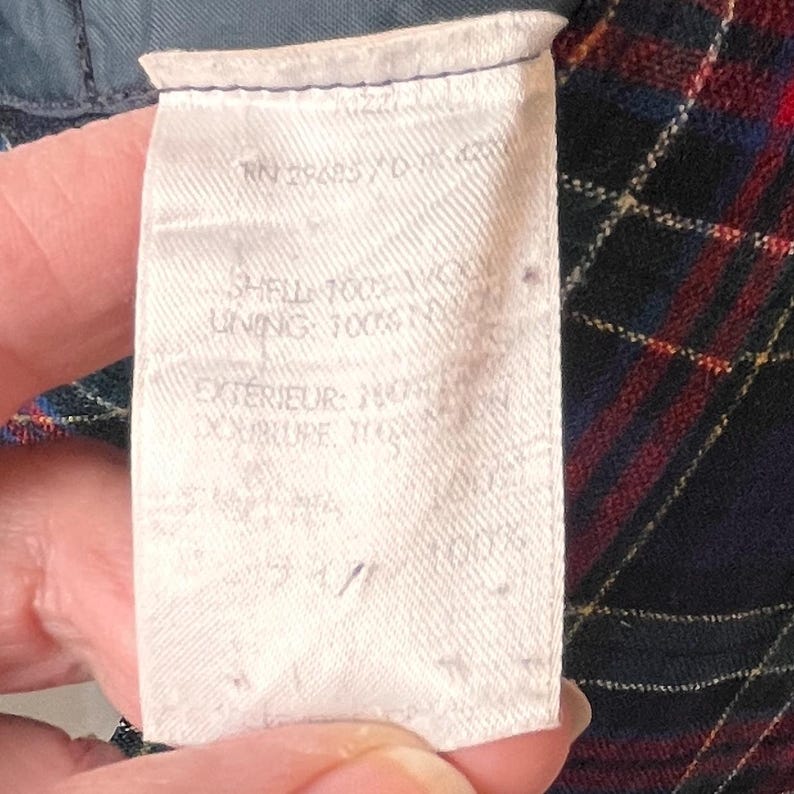 May include: A close-up of a white fabric label with text, held by a hand. The label includes text such as "SHELL 100% WOOL" and "LINING 100% NYLON". The background features a plaid pattern in red, blue, and yellow.